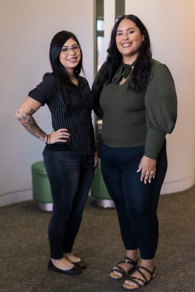 Two women posing indoors, smiling at the camera; one in a striped black top and jeans, the other in an olive-green blouse and dark pants.