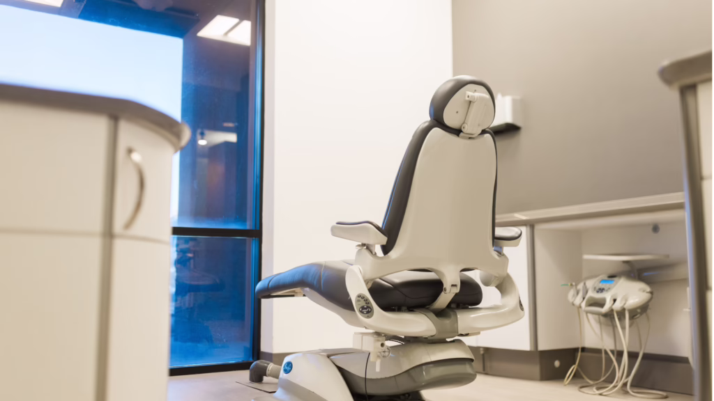 Dental chair in a modern clinical room with beige walls, a blue-tinted window, and medical equipment nearby (treatment station and cords).