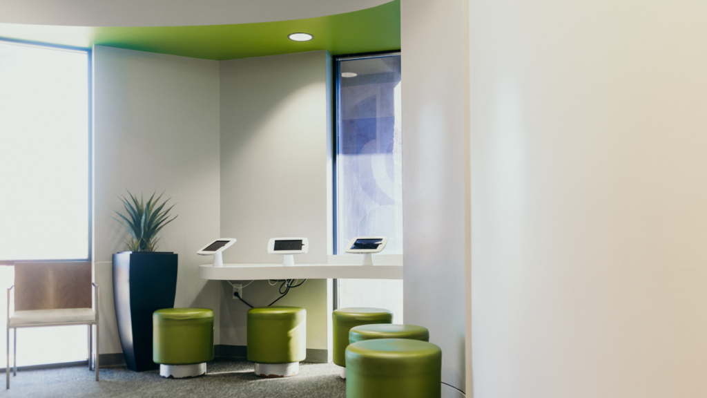 Modern lounge/reception with a white curved counter, three touchscreen kiosks, lime green stools, and a potted plant.