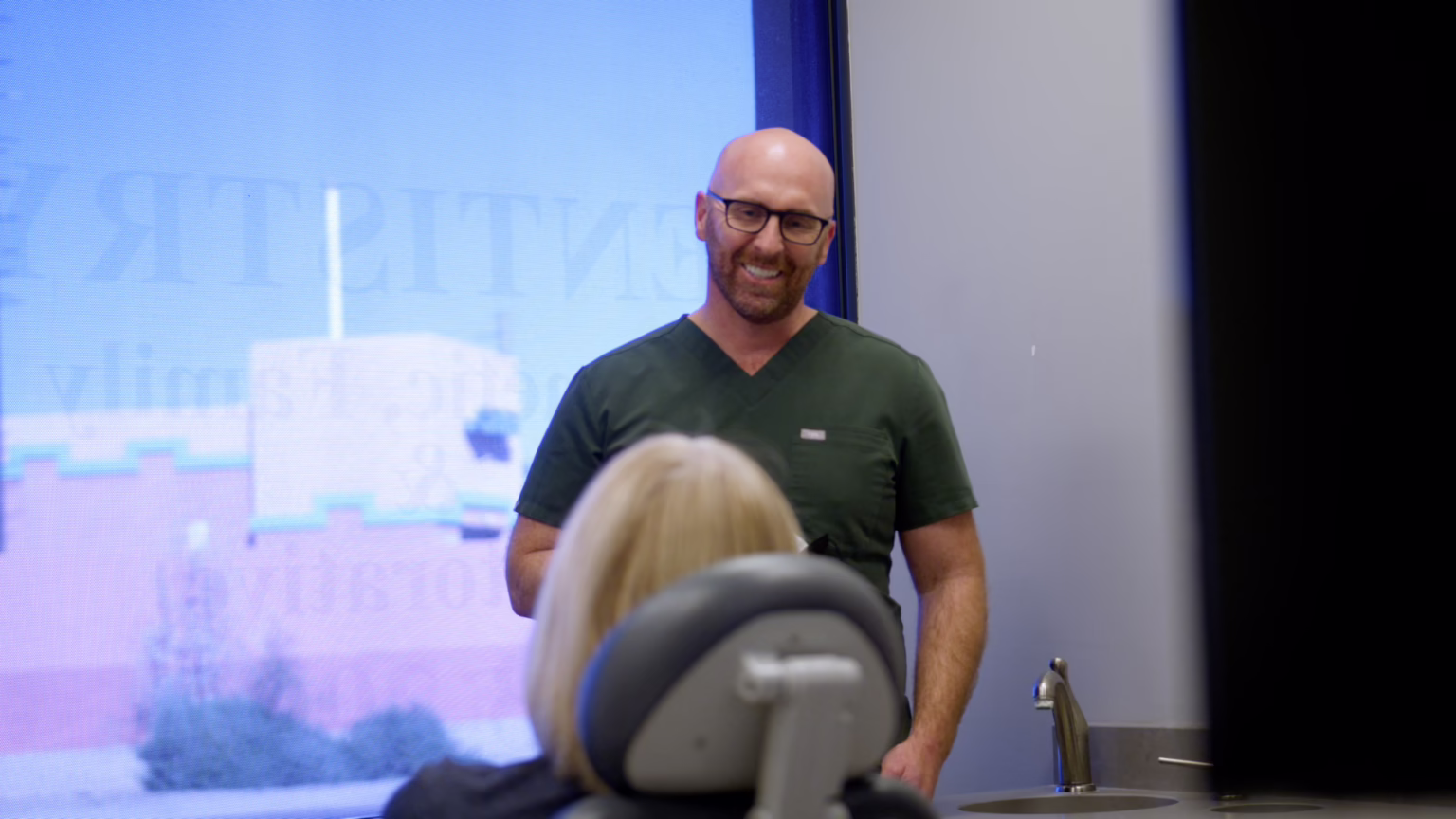 Medical practitioner in green scrubs smiling while speaking to a seated audience in front of a large screen display