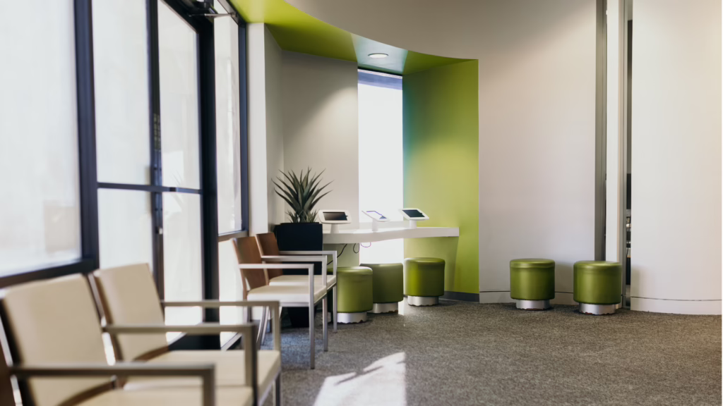Modern waiting area with lime-green accents: row of beige chairs along a windowed wall, a white counter with tablets, and green stools nearby.